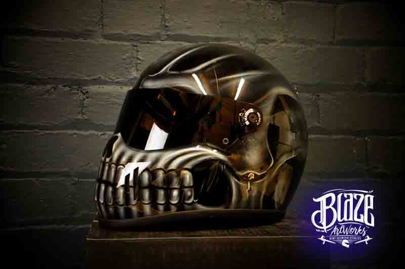 Dark bronze skull motorcycle helmet with metallic airbrushed death head design, detailed teeth and eye sockets against brick wall